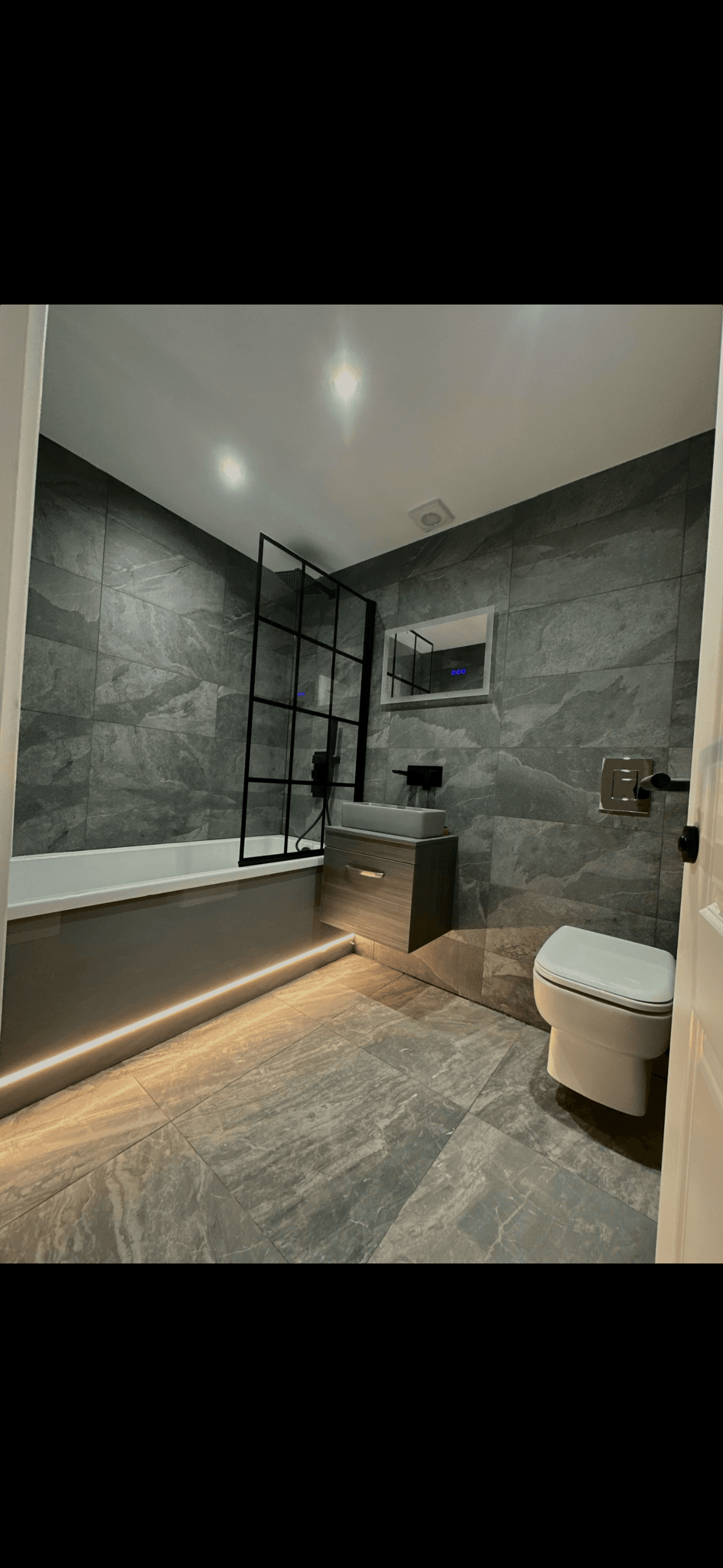Modern bathroom with grey stone tiles, black-framed screen, and warm LED lighting under the bathtub.