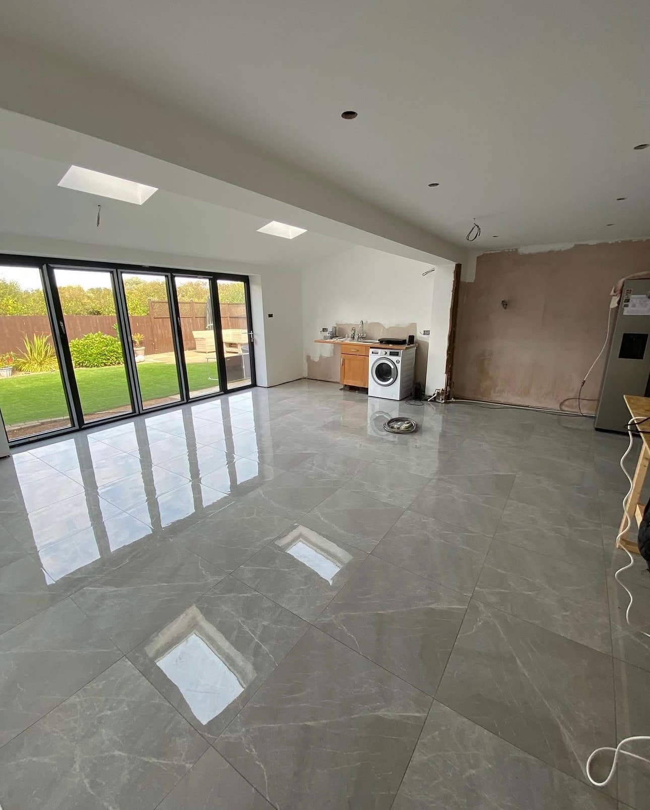 Spacious room under renovation with glossy grey floor tiles, bi-fold doors, and ceiling skylights.