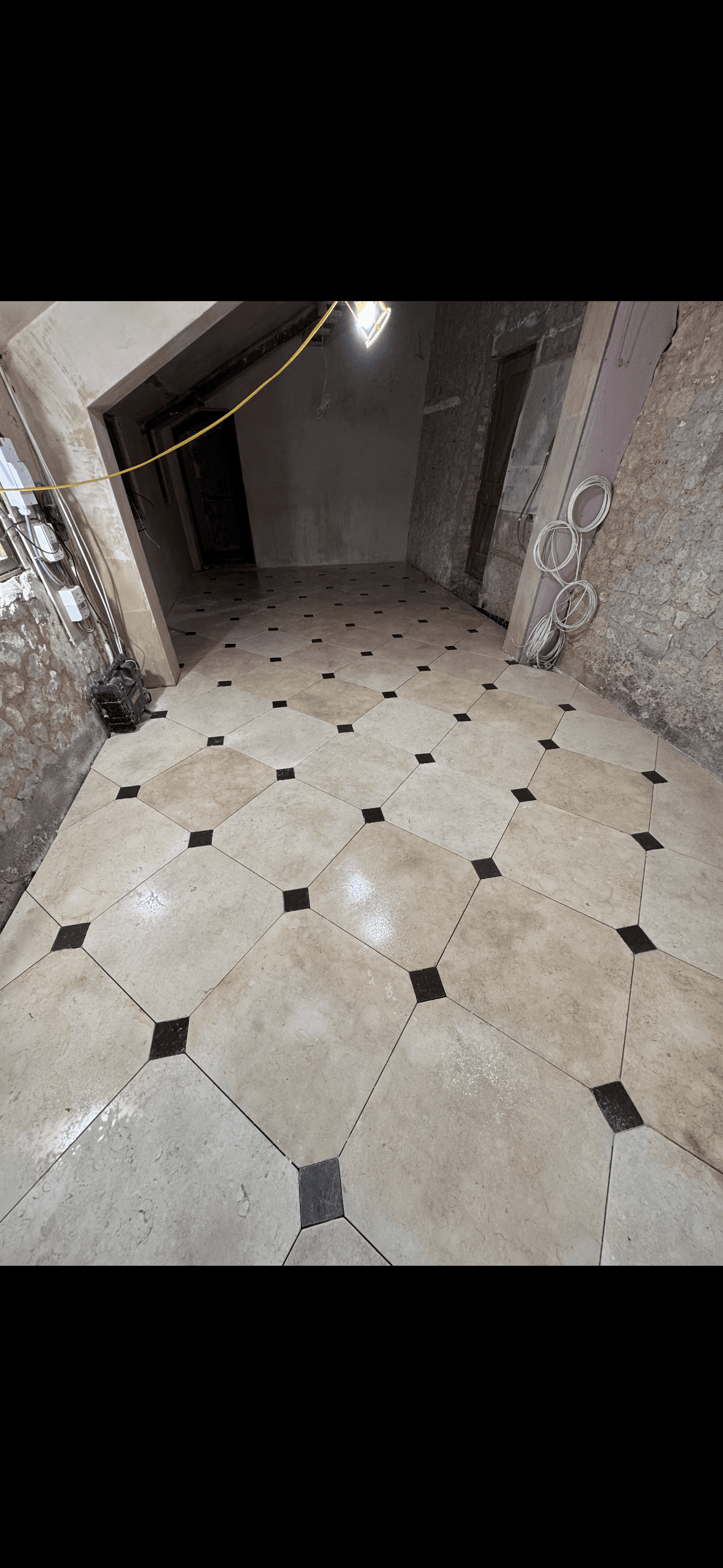 Beige diagonal floor tiles with black accents in a room with unfinished stone walls.