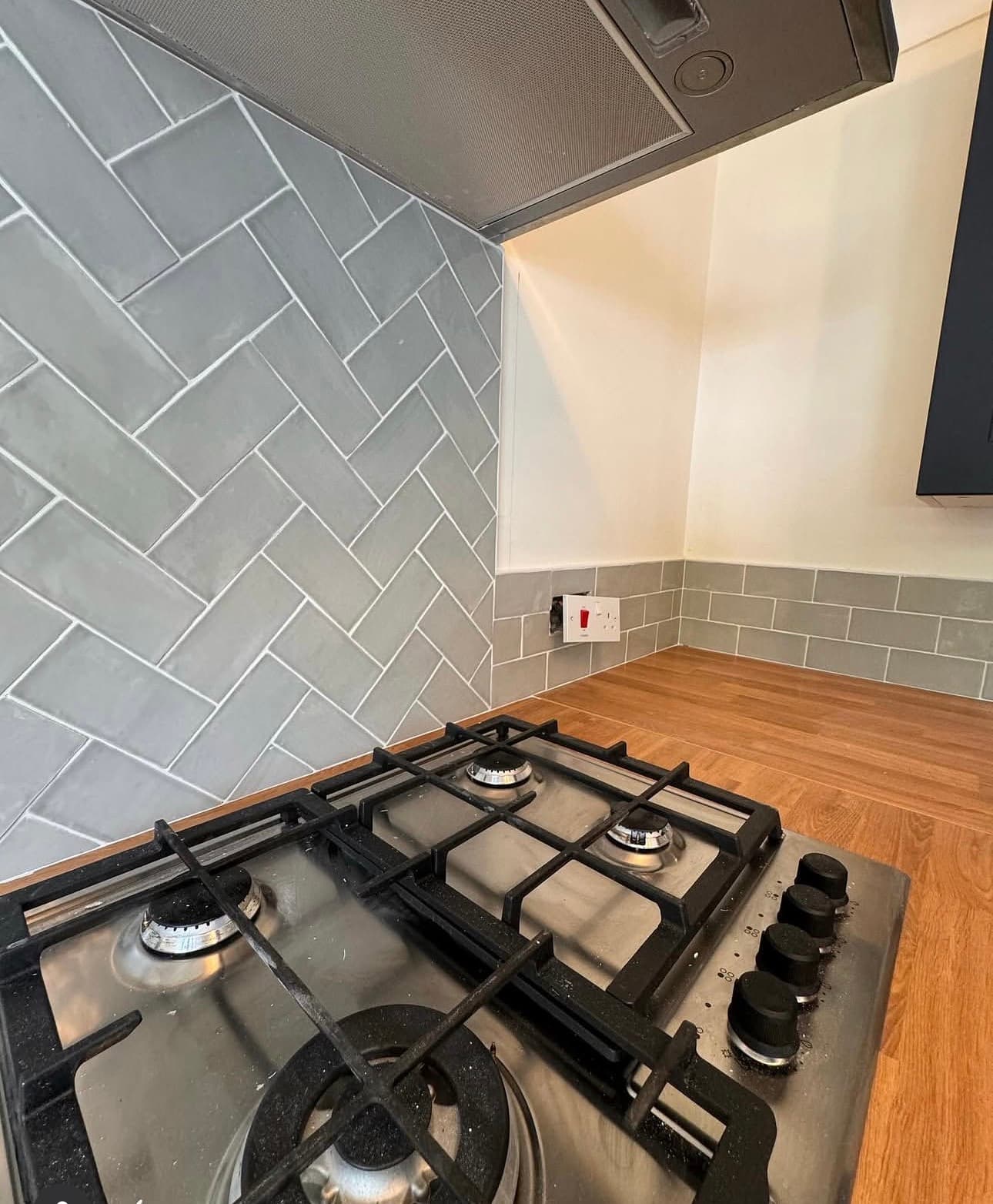 Stainless steel gas stovetop with black grates and a grey herringbone tile backsplash.