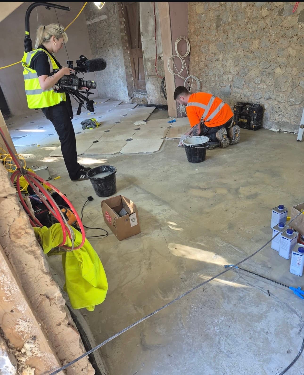 A videographer in a yellow vest films a worker in an orange shirt tiling a floor.