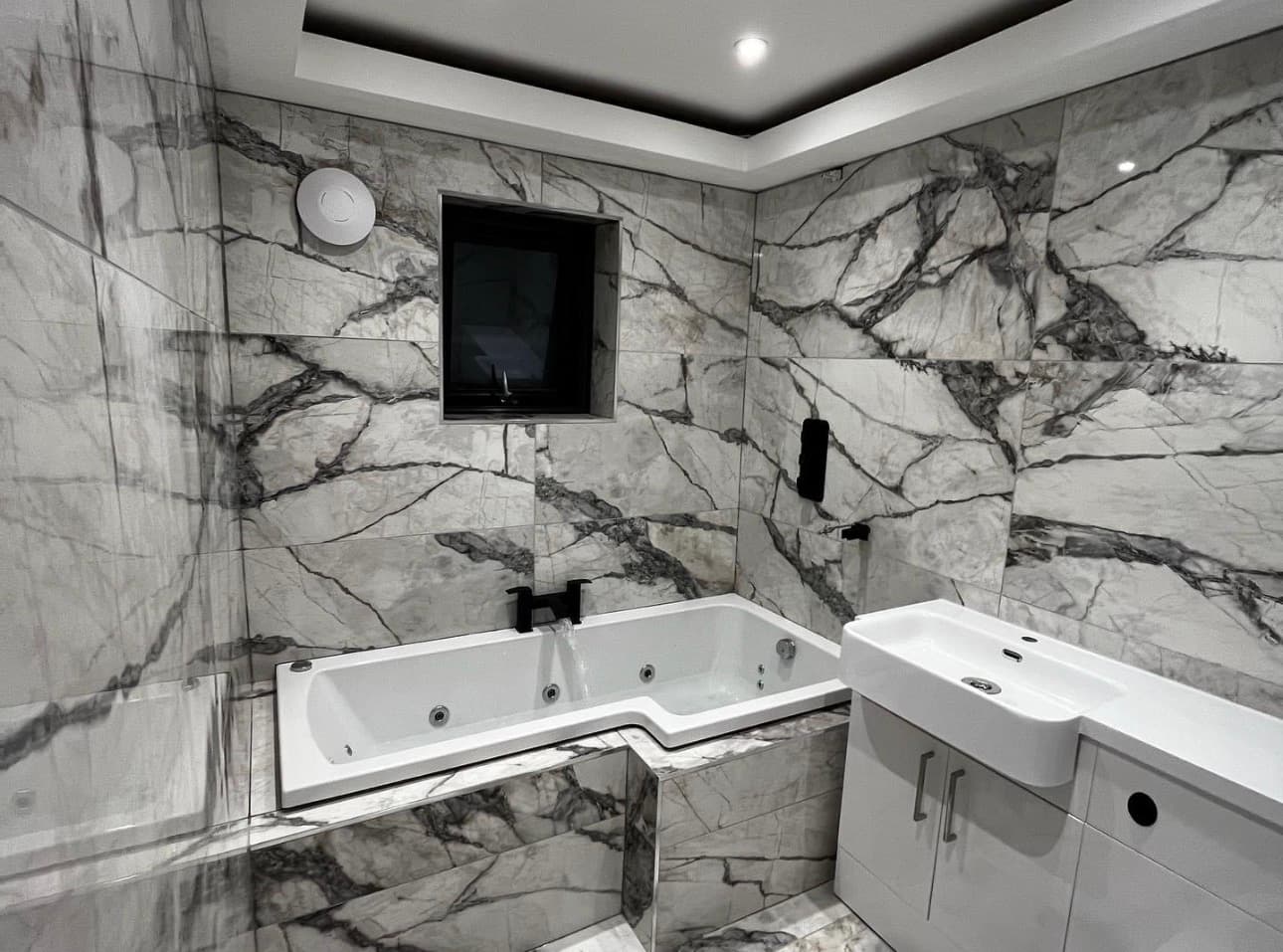 Modern bathroom with grey marble-patterned tiles, a white jetted bathtub, and a white vanity unit.